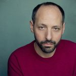 Headshot of Philip Arditti wearing a red top in front of a green background, and looking directly at the camera.