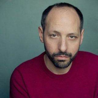 Headshot of Philip Arditti wearing a red top in front of a green background, and looking directly at the camera.