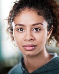 An actor looks to the camera, she wears gold earrings and a blue sweatshirt
