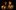 A woman and man stand in the glow of candlelight, the woman's hand touching the man's chin.
