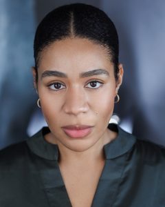 An actor looks to the camera, she has gold earrings and a dark green shirt on
