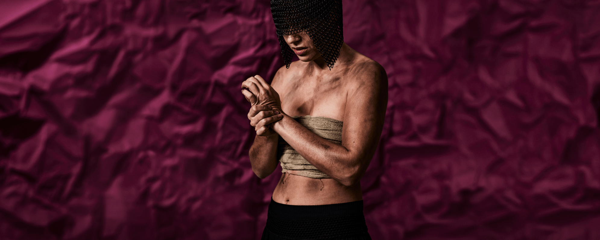 An actor wears a chain metal headpiece which covers their face, they look to the floor and rub their wrist, they are covered in dirt and are barely clothed