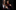 [IMAGE] Two actors stand close behind each other in the dark shadows. One is wearing a three piece suit, and holding a lit tapered candle that drips wax over their hand. The other is wearing a purple knitted jumper, and holds a lit match.