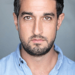 An actor looks into the camera. He has short dark hair and is wearing a blue shirt.
