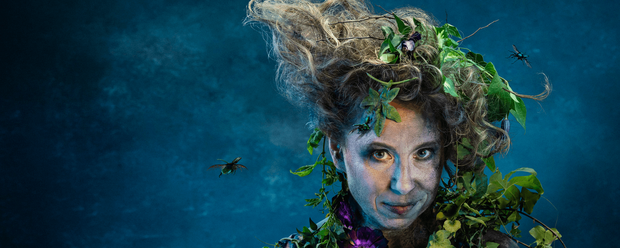 A close up of a smirking woman against a blue backdrop. She is covered in dirt and foliage and has insects flying around her unruly hair.