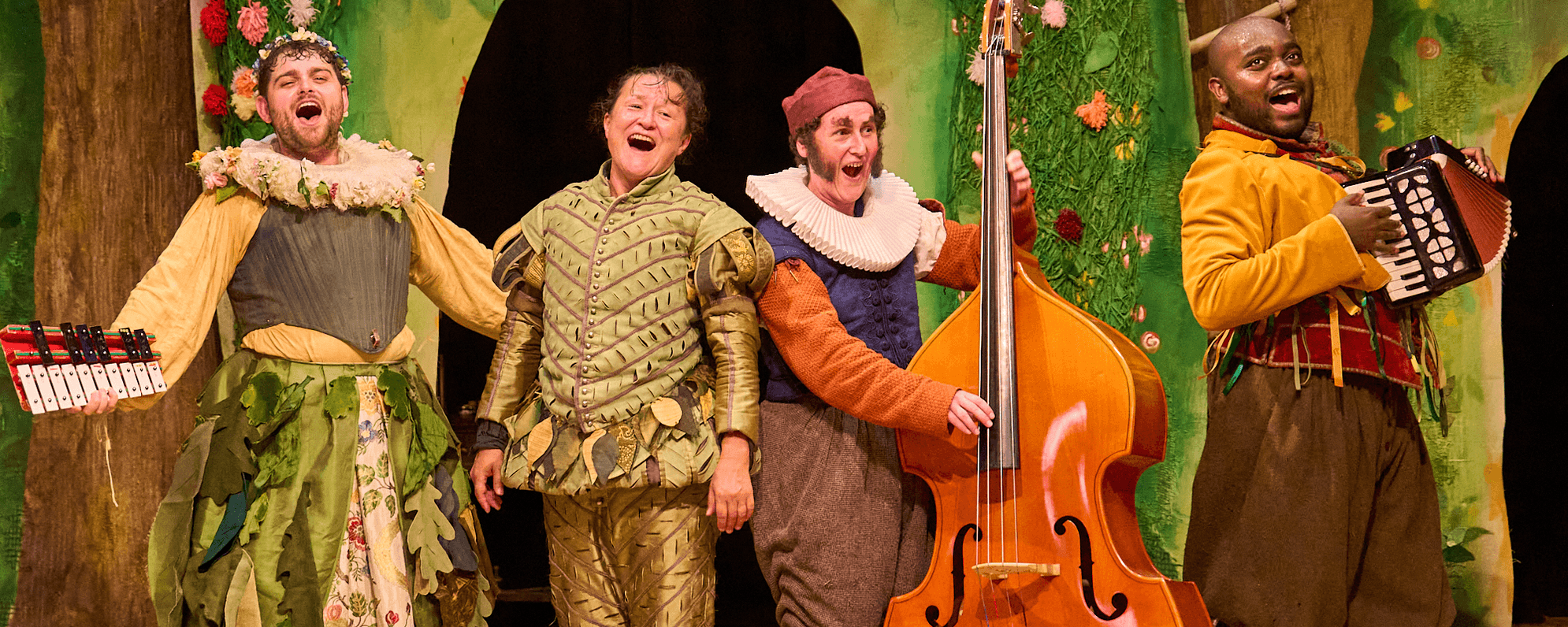 Four actors stand on a stage wearing Elizabethan dress and holding instruments. The set behind them is green, with flowers and trees.