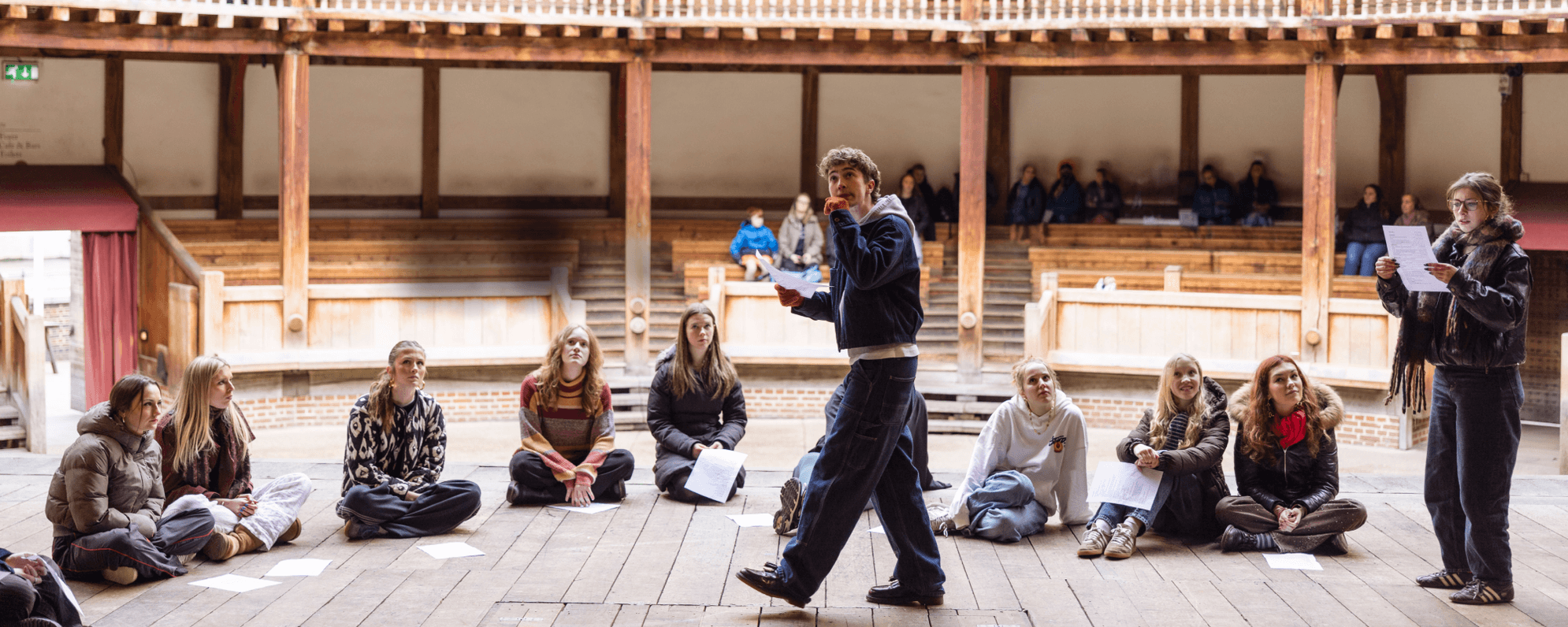 Young people on the stage of a wooden theatre. Some are sitting and some are standing, holding scripts.