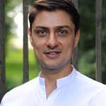 takis looking directly at the camera. takis wears a white shirt, and has short dark hair. takis is outdoors, with trees in the background of the image