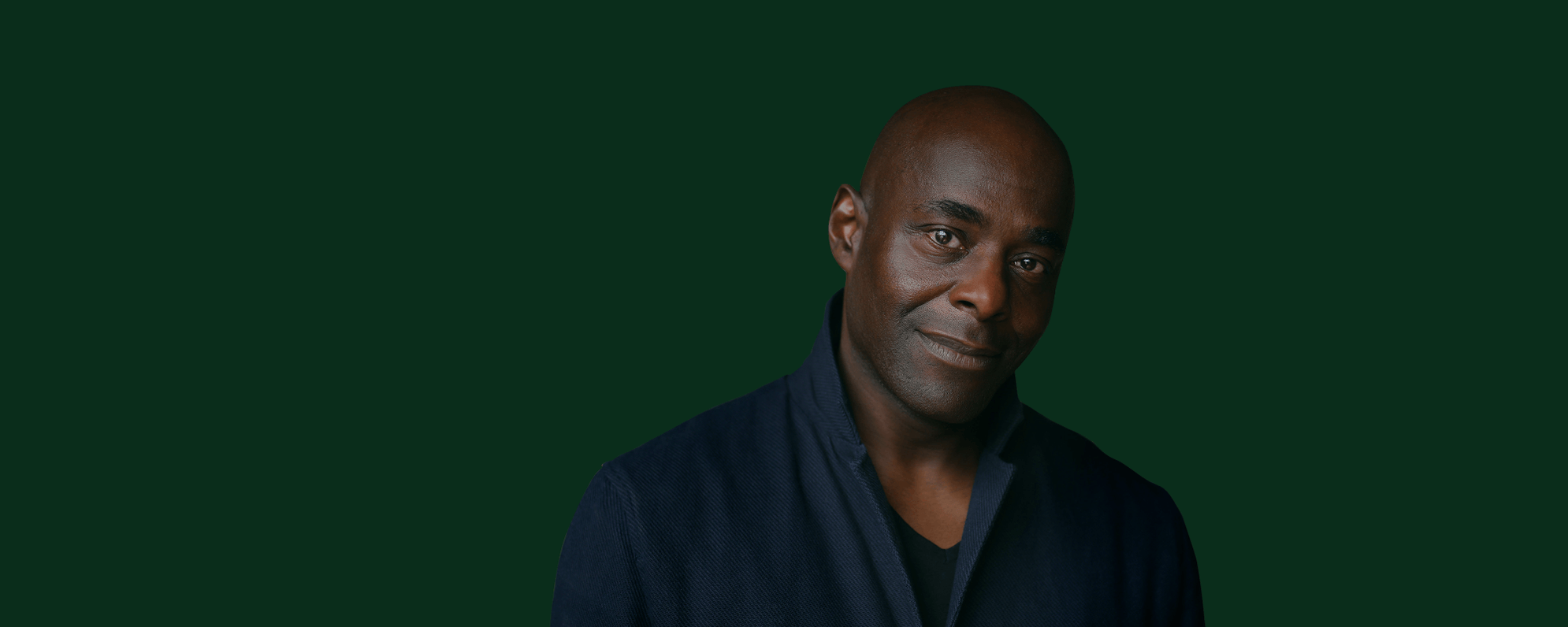 An actor with a bald head and brown eyes wearing a blue jacket looks into the camera, they have a green background behind them