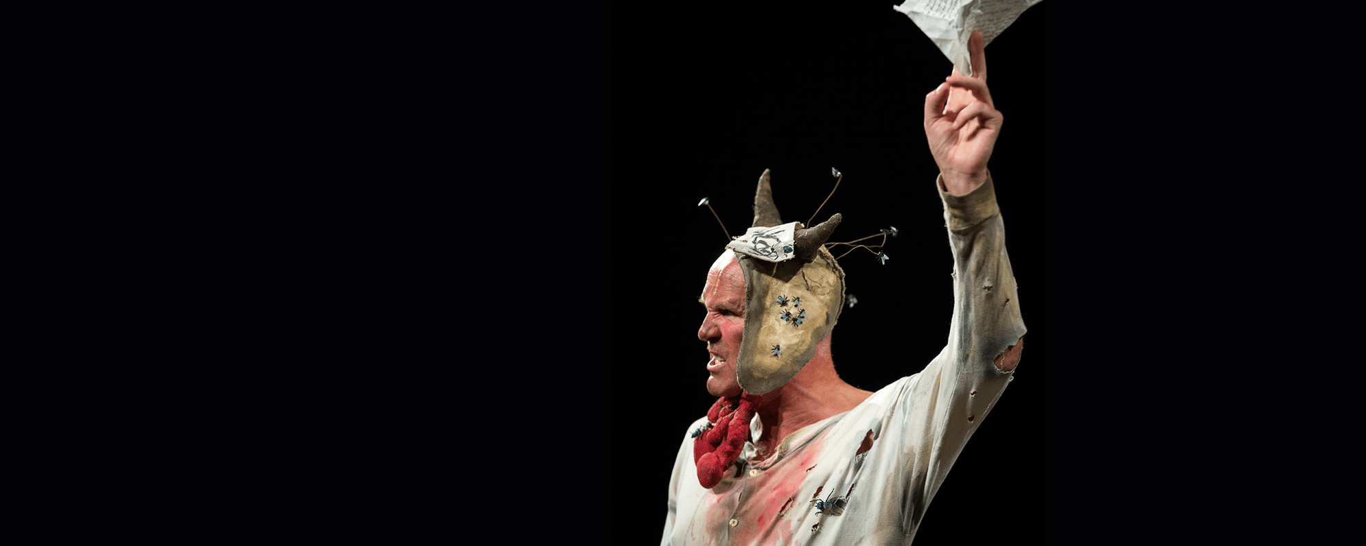 An actor looking angrily off camera, he is wearing a torn white shirt, with a horned leather cap on his head, and holding a scrap of paper aloft in one hand