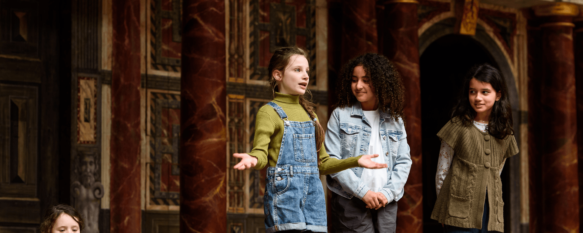 A group of young actors on the Globe Stage, one stepping forward while delivering a speech