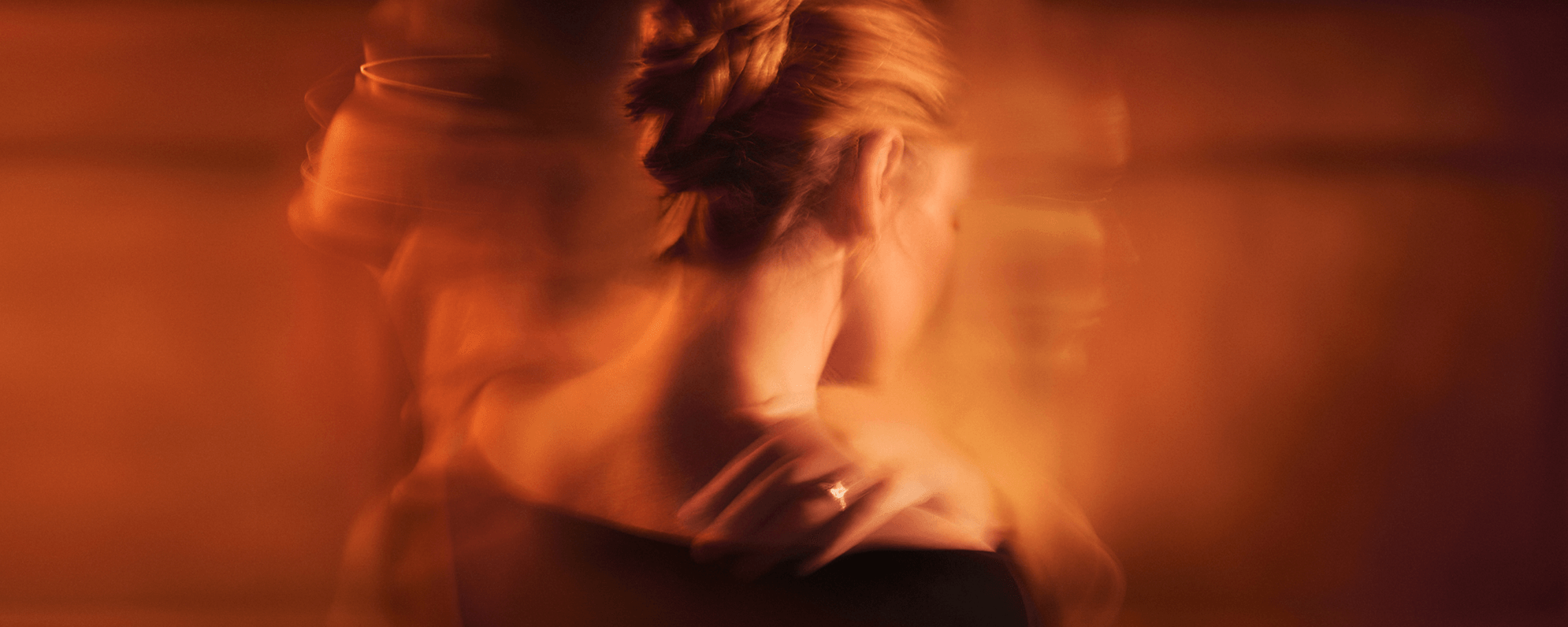An actor facing away from camera, one hand on their shoulder showing a wedding ring. Blurred silhouettes of two other actors are superimposed in the background