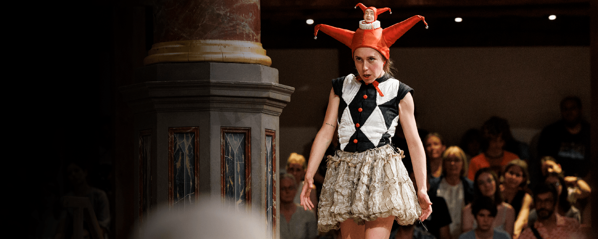 An actor in a jester hat stands on the Globe stage in front of a crowd.