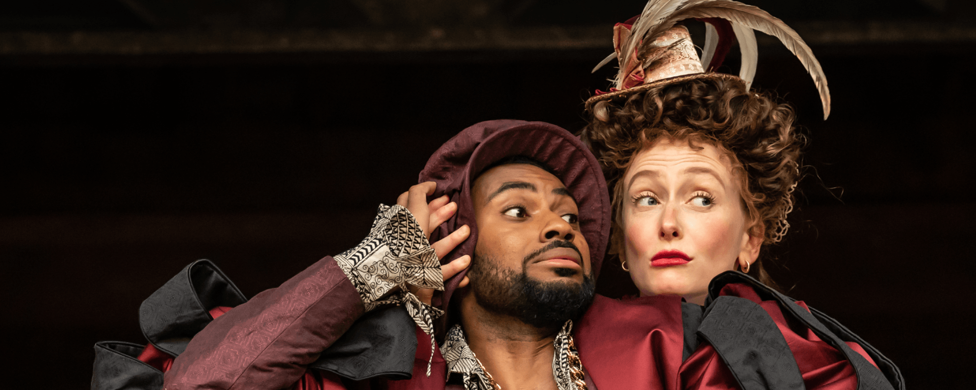 A man and a woman wearing shakespearean dress playfully huddle together on the Globe stage.