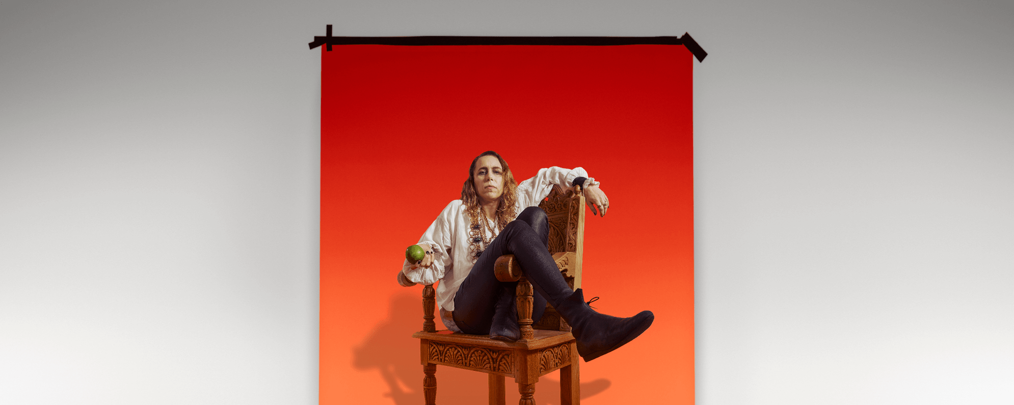 Michelle Terry depicting Richard III sat in a wooden thrown holding an apple in front of a red-orange background.