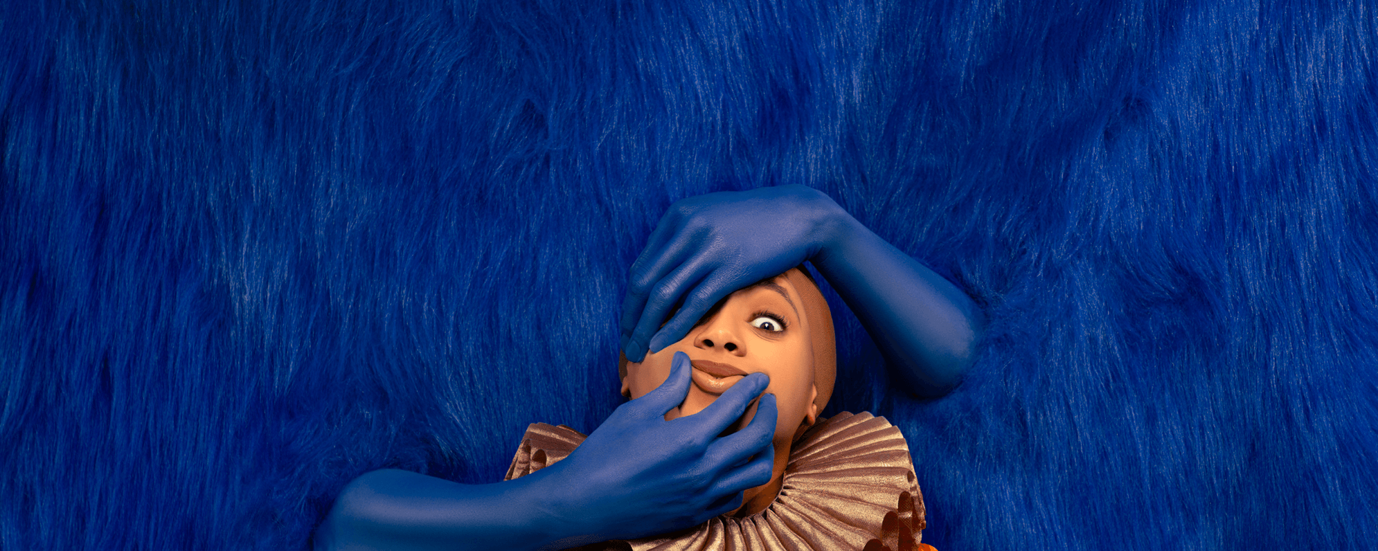 Woman in a ruff stands in front of a blue fluffy background through which blue arms are protruding to manipulate her face.