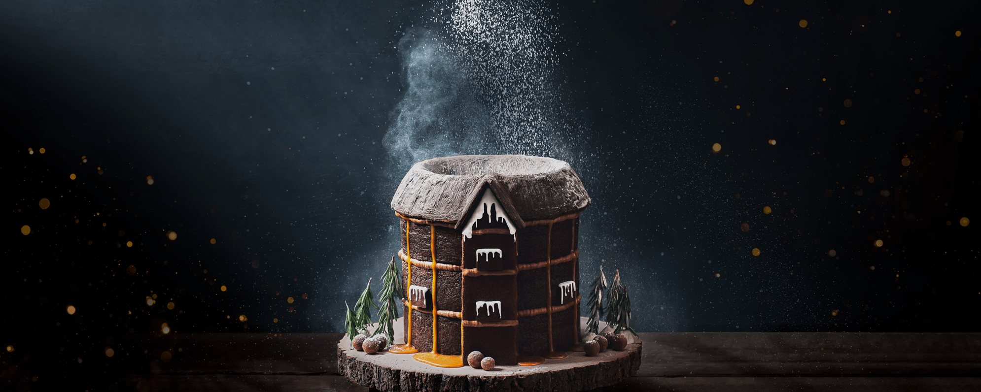'Hansel and Gretel' show artwork depicts a replica of the Globe made from gingerbread cake sits on a log slice in the centre of the image, surrounded by truffles and rosemary sprigs that imitate trees and boulders. White icing mimics icicles, and some orange icing drips down the walls. Powdered sugar drifts onto the platter like snow.