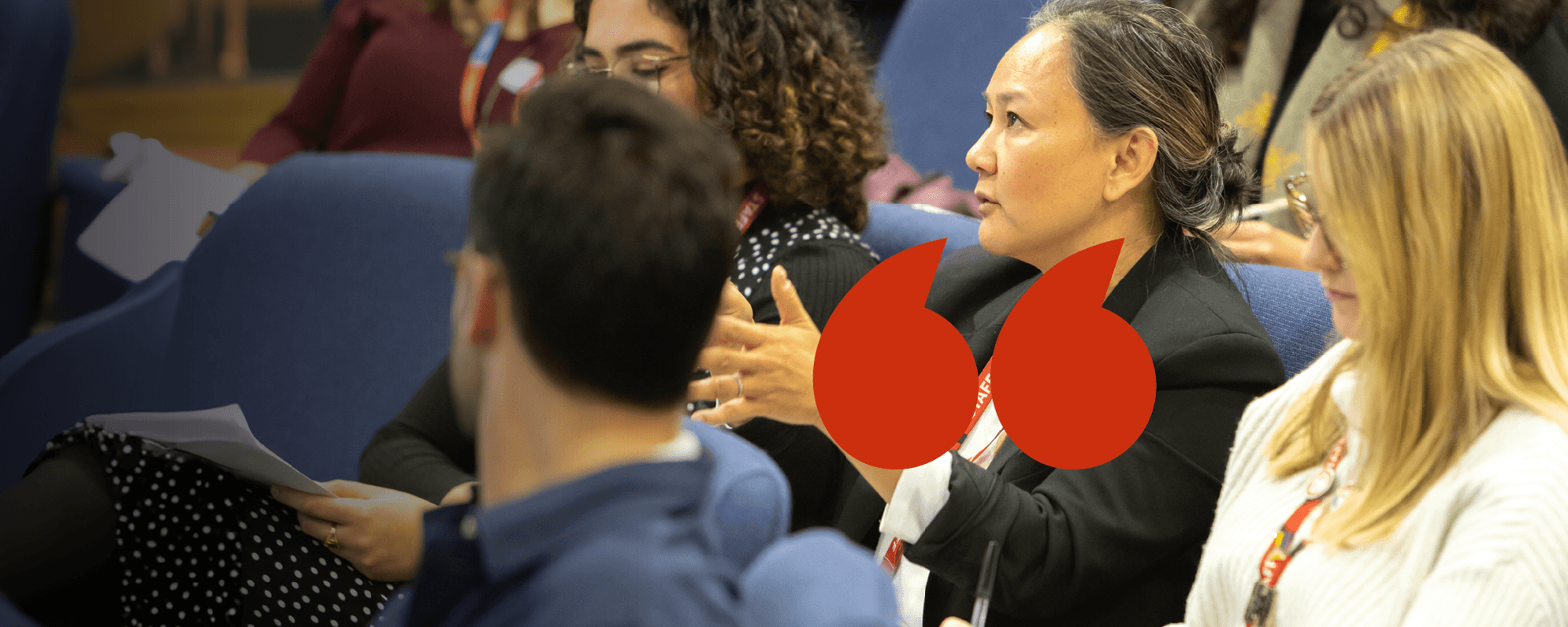 An audience member in a lecture hall and addresses someone. A red apostrophe motif is overlaid.