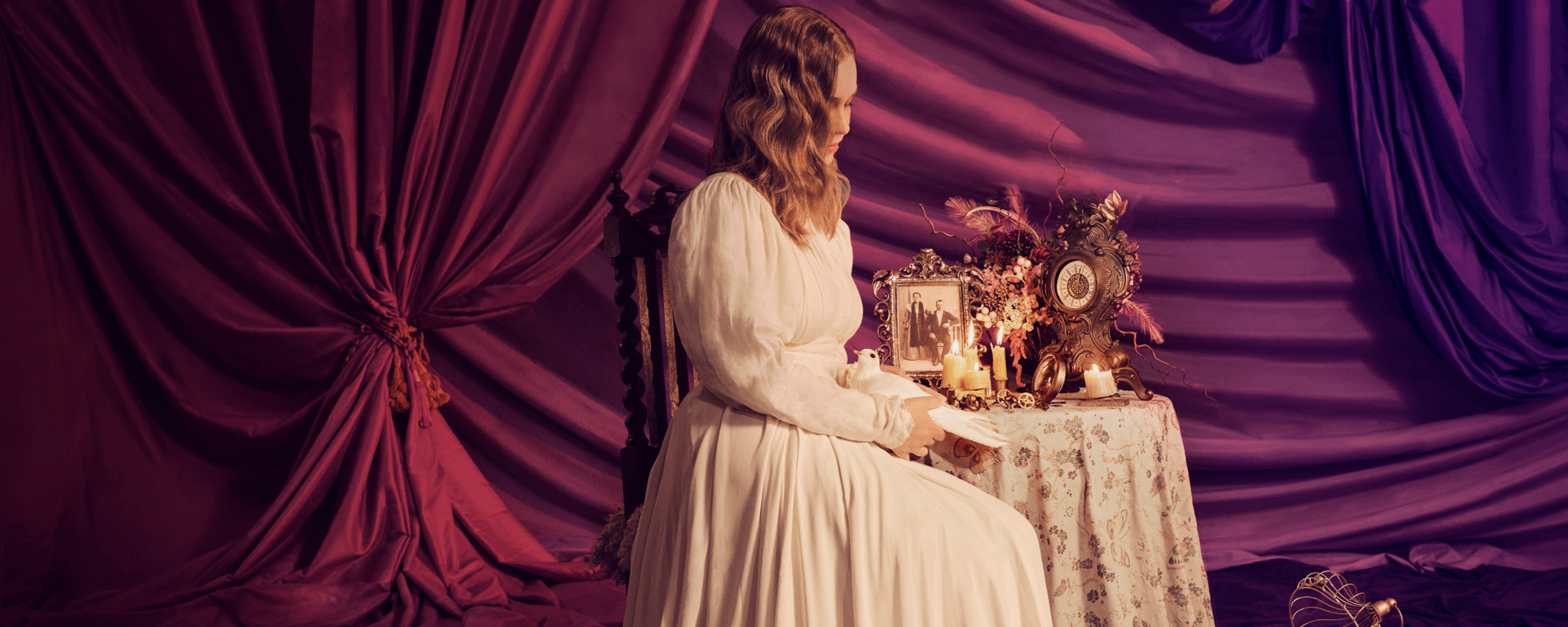 'Three Sisters' show artwork depicts a woman in a white long-sleeved dress holding a white dove. She sits in a wooden chair in front of a small table against background of purple curtains. The table is covered by a patterned cloth, an ornate clock piece, a black-and-white framed photograph, and candles.