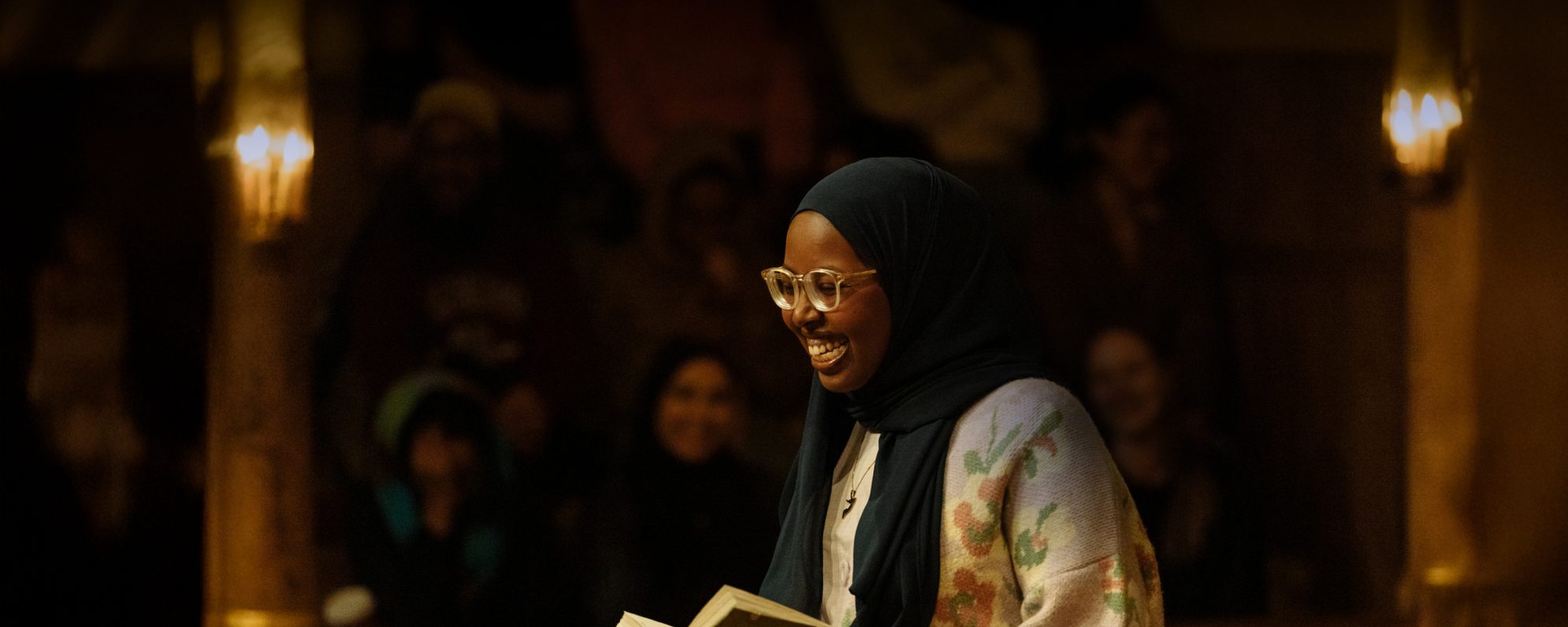 A person wearing a headscarf and glasses smiles in a dimly lit room with audience members blurred in the background.