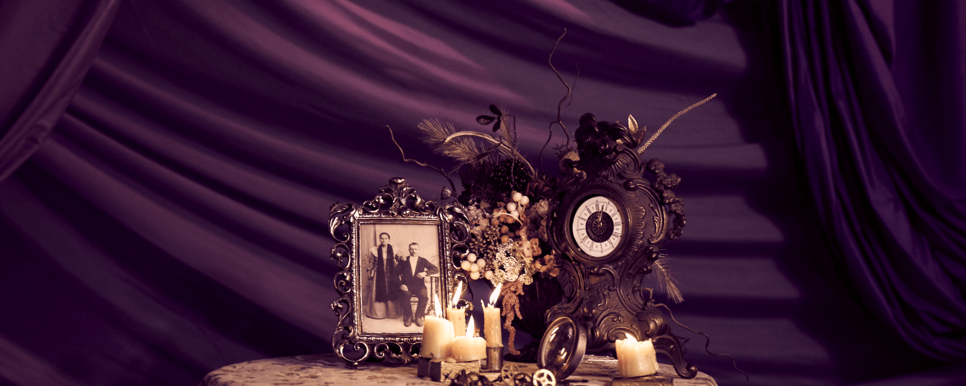 There is a purple curtain in the background and a small table. On the table there are 19th Century objects including an old photograph, a clock, and some candles.