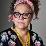 Naomi Wirthner looking directly at the camera. Naomi is wearing a patterned shirt, necklaces, and a pink cloth headband.