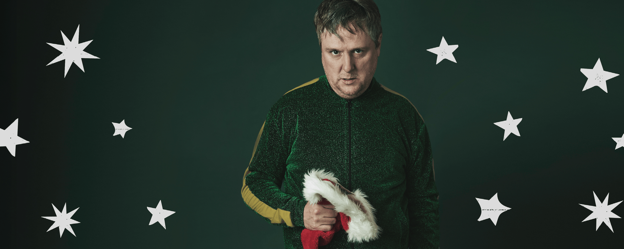 Tim Key, clutching a Santa hat, in a sparkly green tracksuit, against a green background with white stars