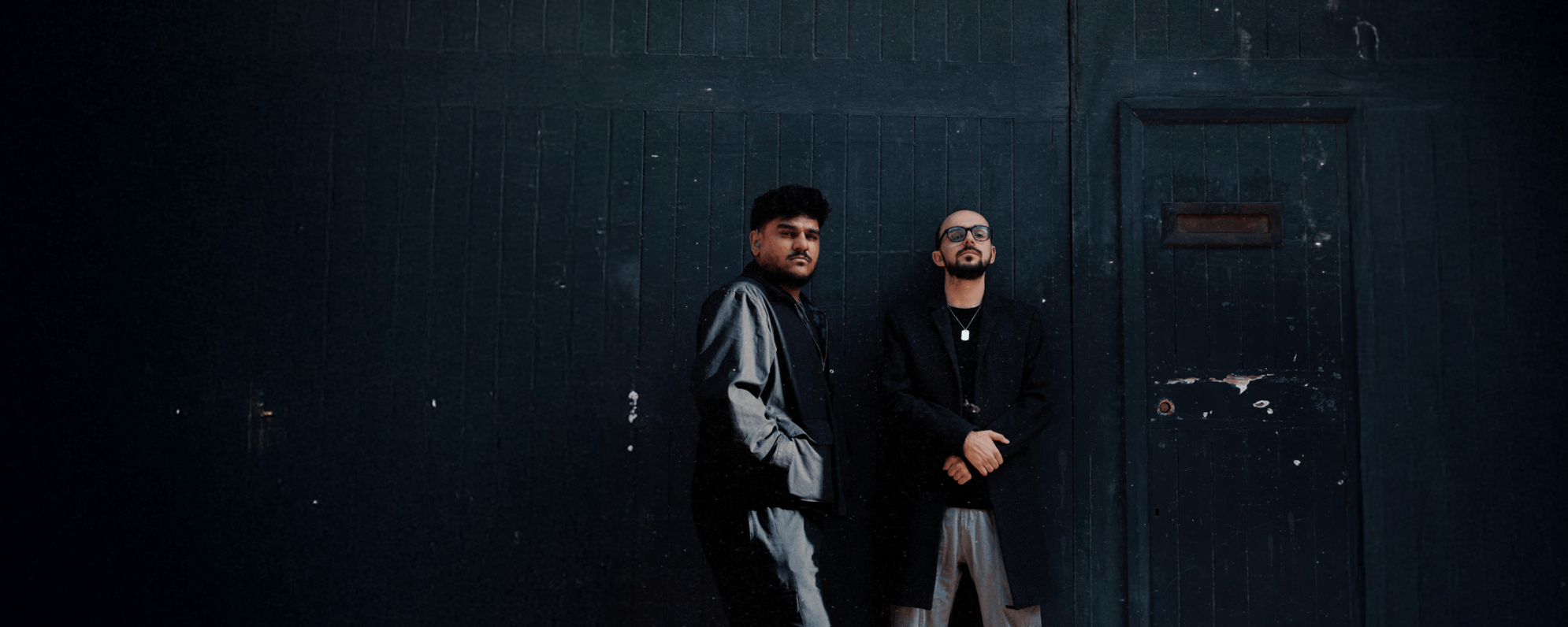 Nadeem Islam and William Grint stand before a dark wooden background, both looking directly down at the camera.