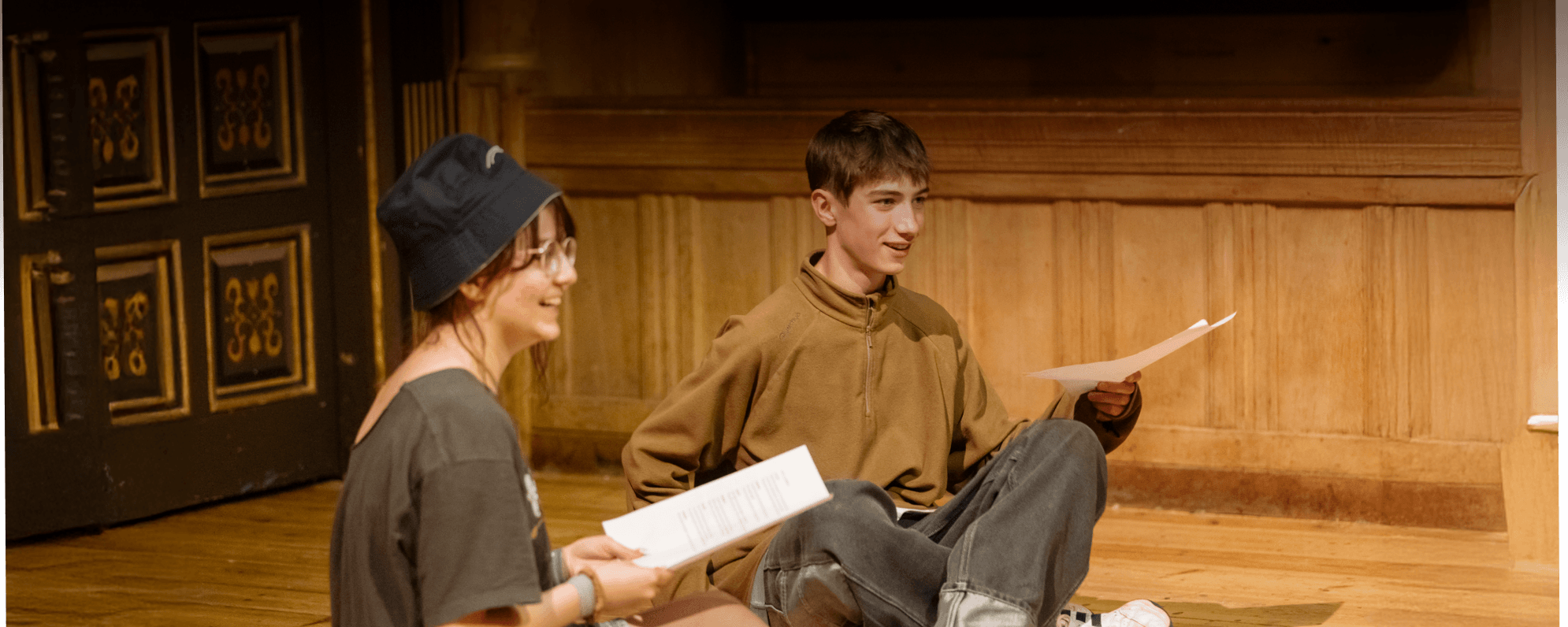 Two young people sat cross-legged on the Sam Wanamaker Playhouse stage with scripts