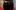 A black fluffy cat stands on a table with a red tablecloth, looking up at the camera.