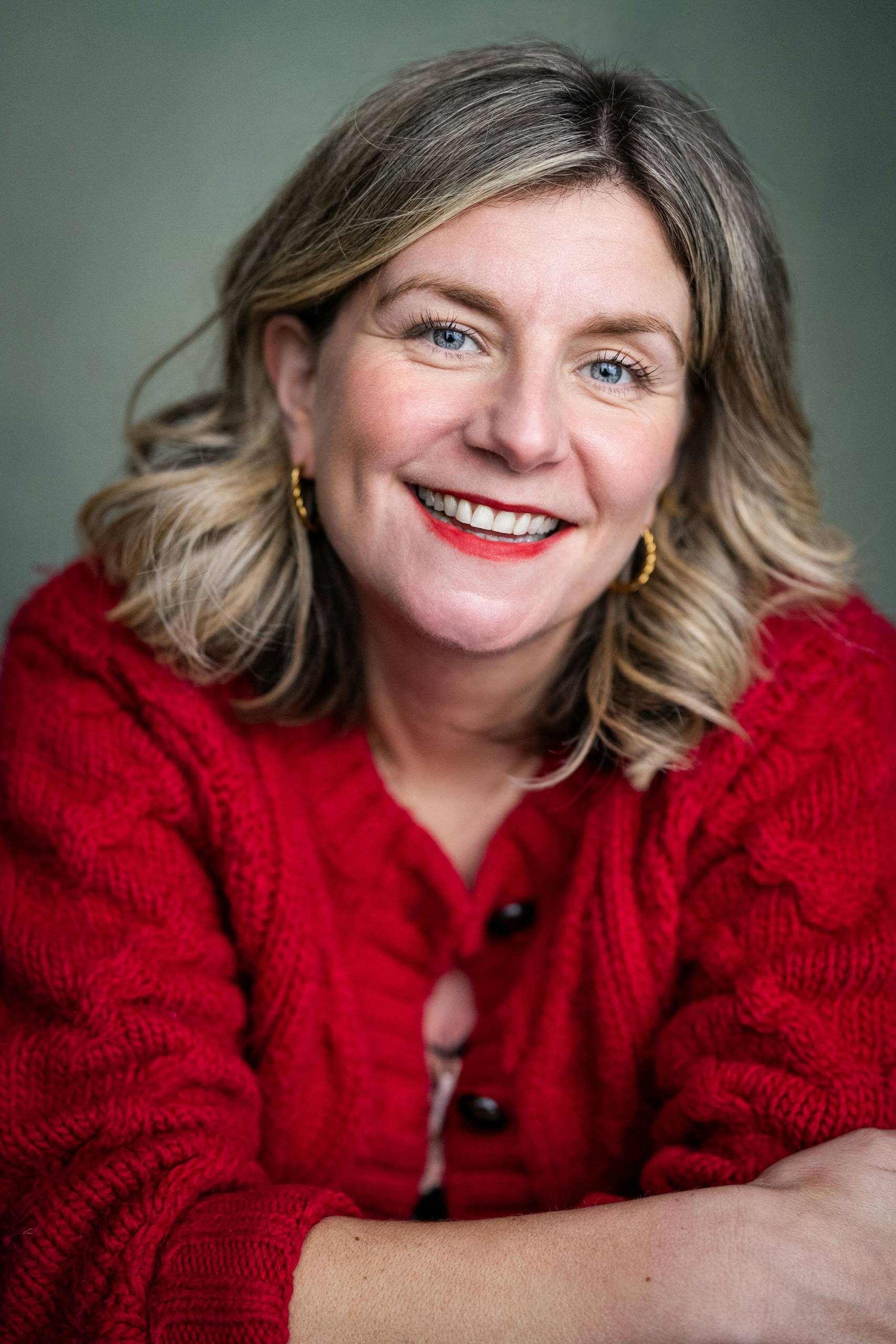 Anna Jordan smiling directly at the camera. They have shoulder-length, blonde curled hair and are wearing a red knitted cardigan.