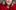Anna Jordan smiling directly at the camera. They have shoulder-length, blonde curled hair and are wearing a red knitted cardigan.