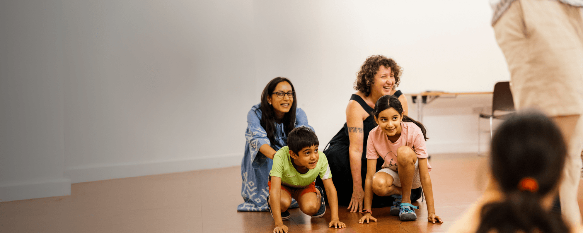 Two children kneel on the floor and two adults kneel behind them, laughing whilst taking part in a family workshop.