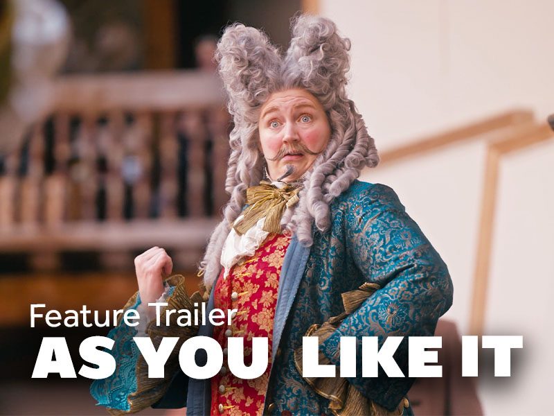 Feature Trailer. As You Like It. A white woman wearing a silver wig and moustache in a blue and red Baroque men's outfit pulls an exaggerated expression and gestures behind her with her right thumb.