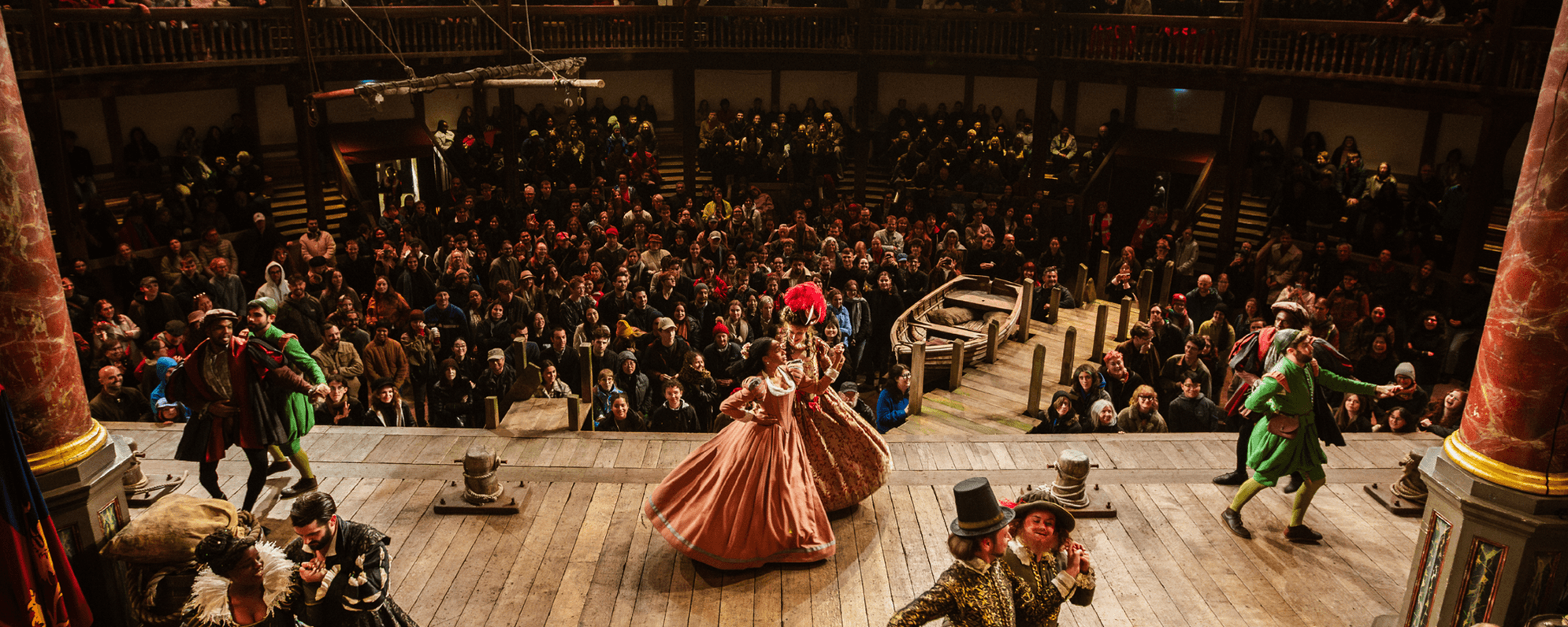 An audience watch at nighttime while performers wearing historical costumes dance in pairs around a wooden stage.