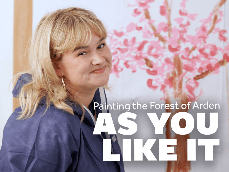 Painting the Forest of Arden. AS YOU LIKE IT. Lola Shalam smiles at the camera beside a painting of a tree.