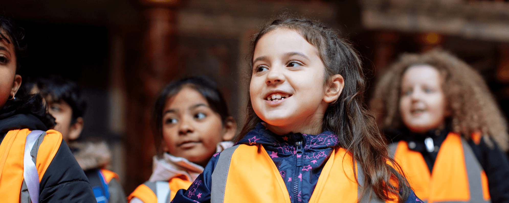 A group of primary school children in the Globe Theatre. They are wearing orange hi-ves vests and raincoats.
