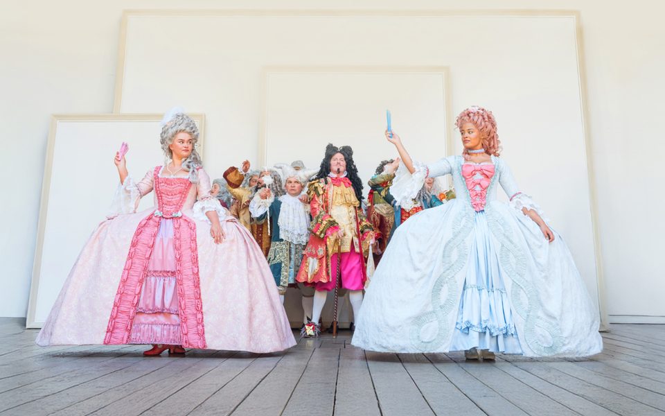 A White woman with dark brown eyes, wearing a large grey wig and a baroque era pink dress and a mixed race woman wearing a pink wig and a blue baroque dress, stand on stage holding fans and looking at each other. Behind them are a group of people and a white background.