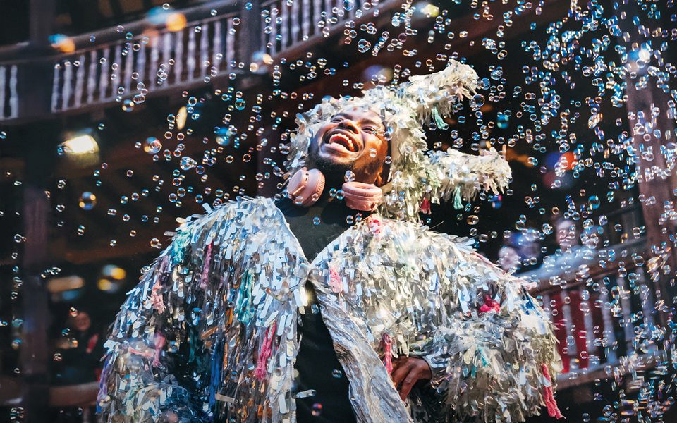 A tall dark-skinned British-Gambian man with short Afro hair wears a silver tassel jacket and silver donkey ears. He stands surrounded by bubbles.