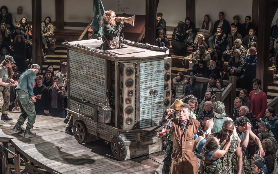 In a round theatre with wooden seating galleries, an audience watches as a parade of people wearing camouflage dance around a large blue cart. Standing in the cart is a white woman with long curly hair, speaking into a megaphone.