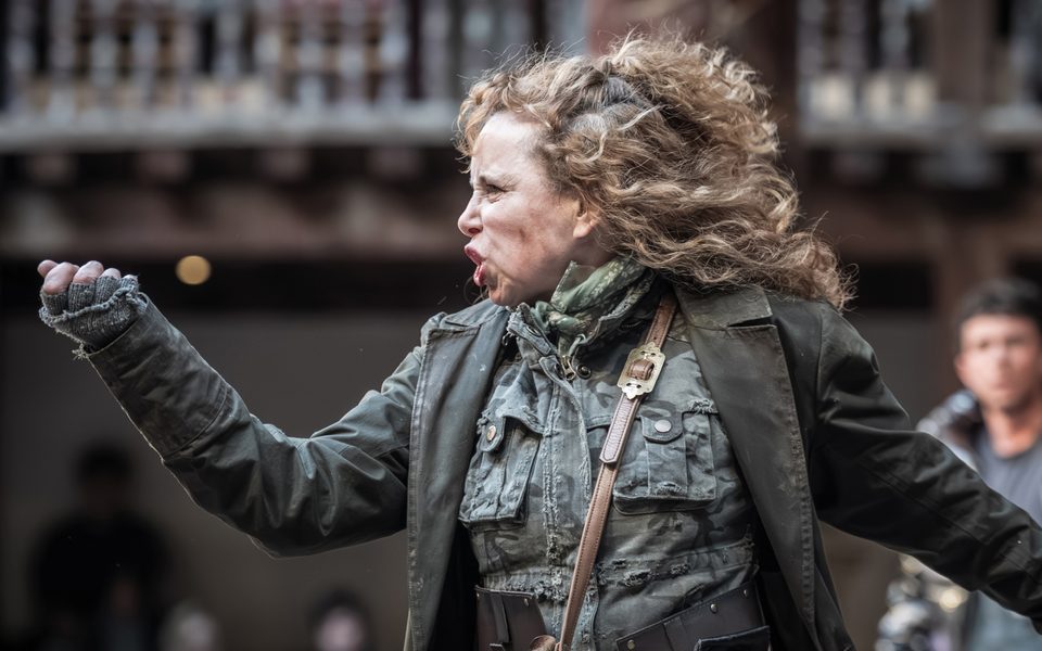 A white woman with curly blonde hair, wearing a khaki jumpsuit and green coat, points a knife in front of her and contorts her face. A wooden theatre can be seen behind her.