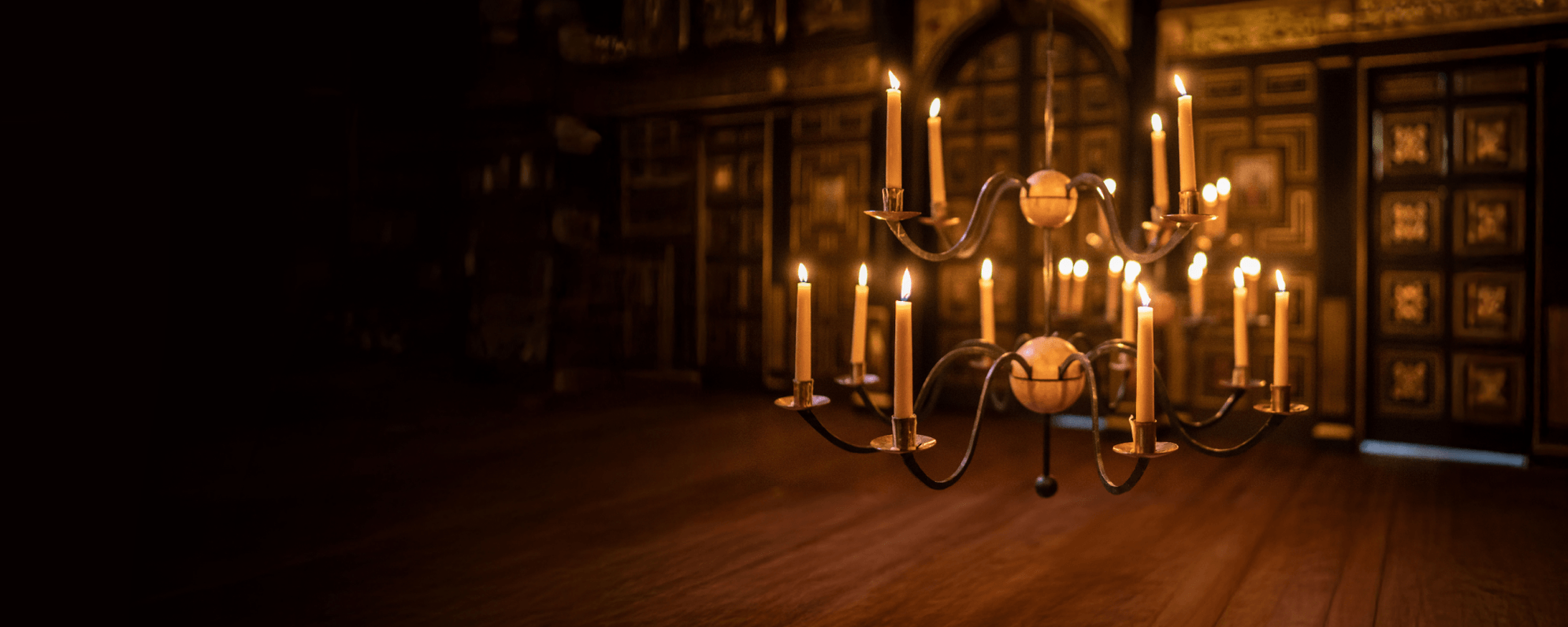 A chandelier of lit candlesticks hangs down from above. It illuminates a wooden floor beneath and a black and gold embellished wall behind.