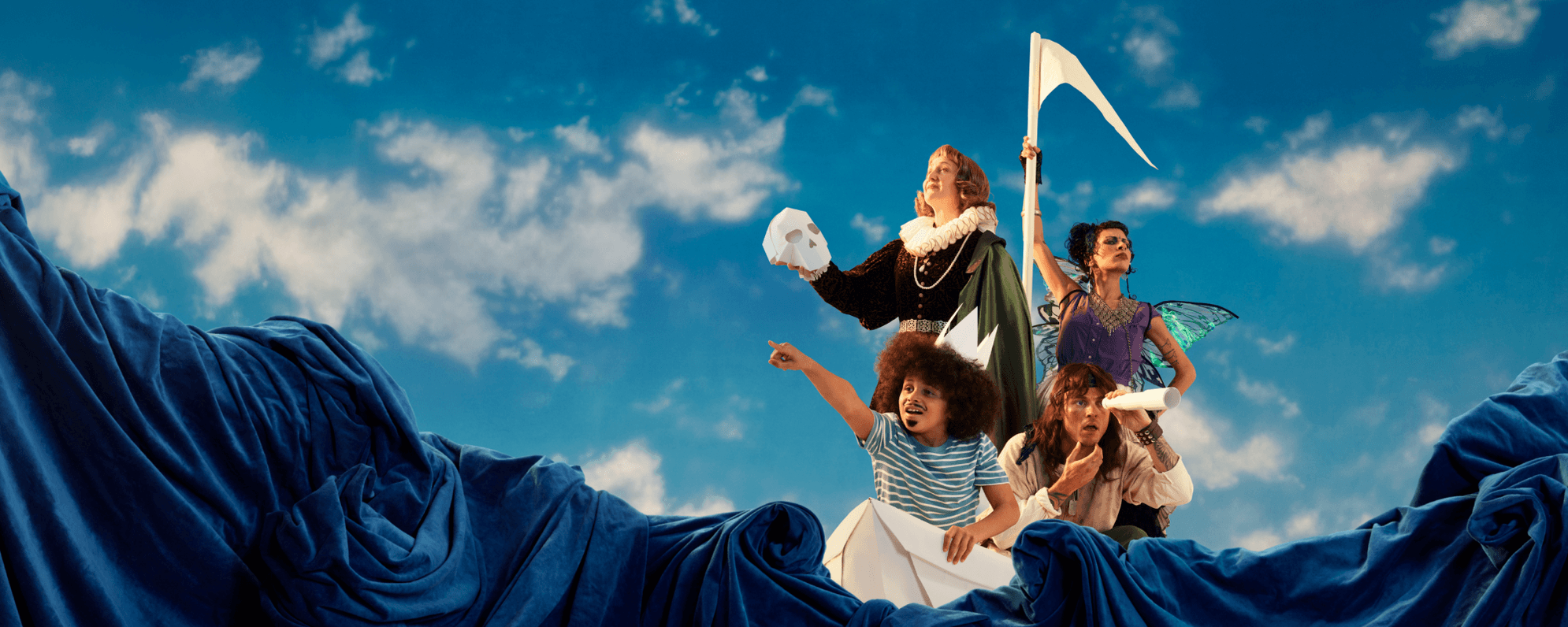 Four people are aboard a giant paper boat against a bright blue, cloudy sky and a sea of dark blue material. Two are standing; one in Shakespearean dress holding a paper skull, another in a purple fairy costume holding a paper mast. The other two are crouched, with one wearing a paper hat, pointing out to sea, and another looking through a paper telescope, scratching their chin.