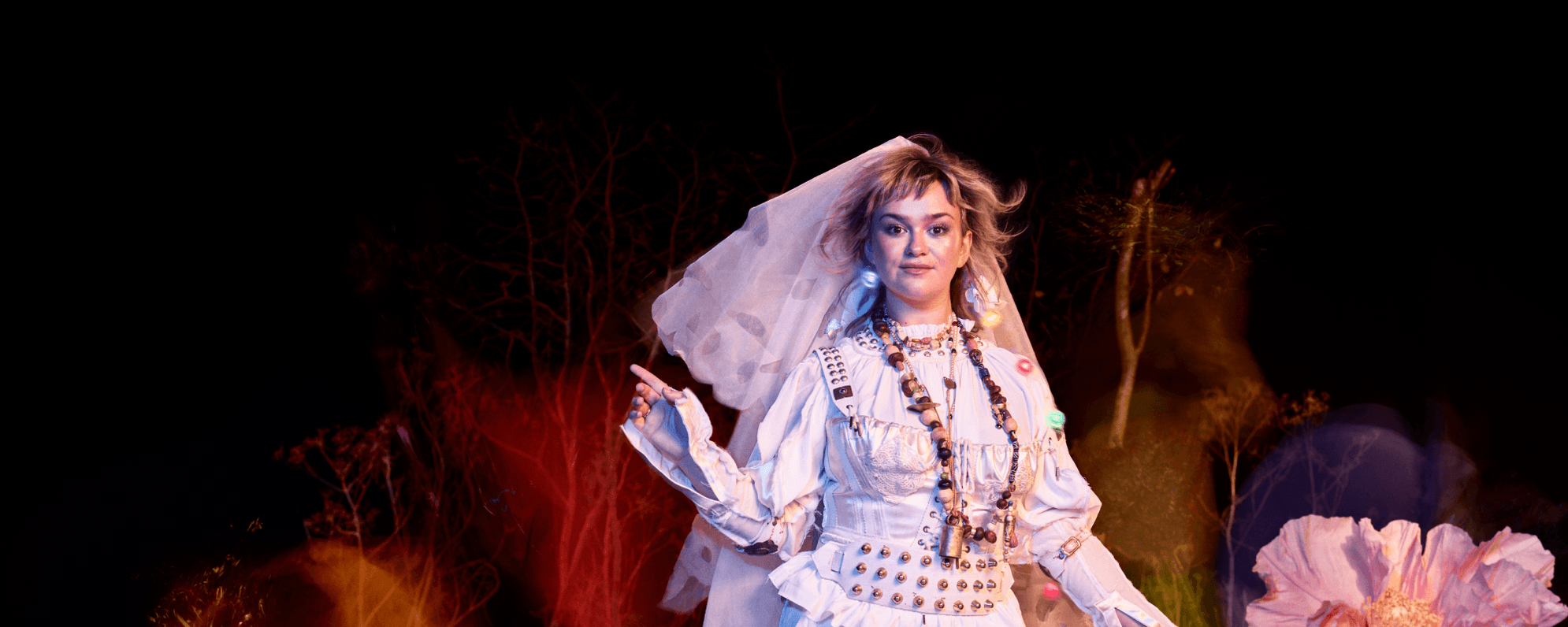 A person wearing a white shirt and a pinafore dress with a veil, smiles gently directly into the camera, whilst pointing up with their right hand. They have blonde hair and brown eyes and wear a large bead necklace in various shades of brown. Small orbs of colour appear around them. In the background, various trees are overshadowed by tints of red, yellow and purple.