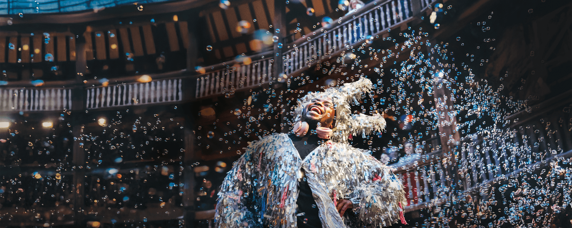 A tall dark-skinned British-Gambian man with short Afro hair wears a silver tassel jacket and silver donkey ears. He stands surrounded by bubbles.