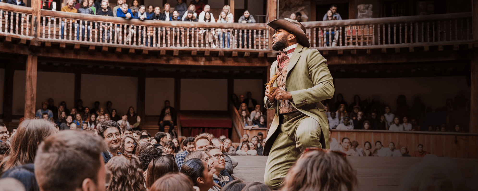 An actor wearing a green suit and cowboy hat kneels down at the front of a wooden stage. Audience members stand below the stage, looking up at the actor. In the background, audience members sitting in wooden seating galleries watch on.