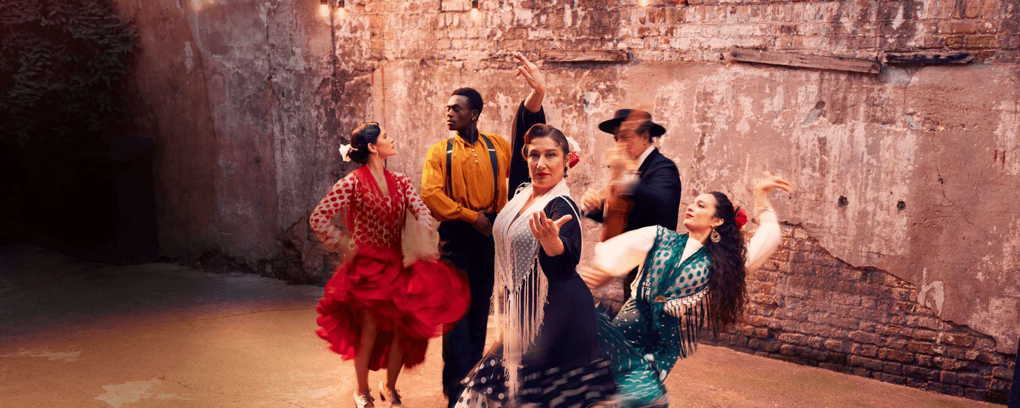 A group of flamenco performers are captured in motion against a plain brick wall. Four of the group, wearing traditional colourful flamenco costumes, take strong dance positions. A fifth person wearing a dark suit stands playing a guitar, which is blurred through the capture of movement.