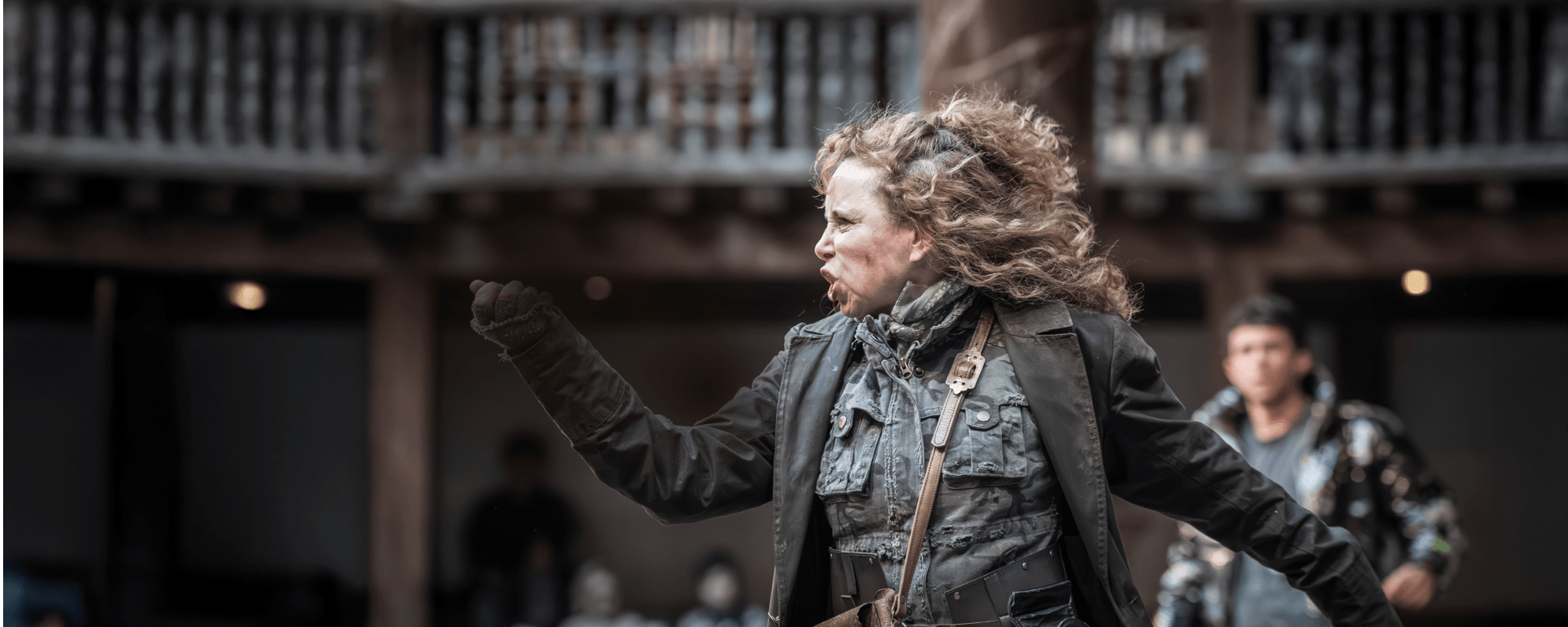 A white woman with curly blonde hair, wearing a khaki jumpsuit and green coat, points a knife in front of her and contorts her face. A wooden theatre can be seen behind her.