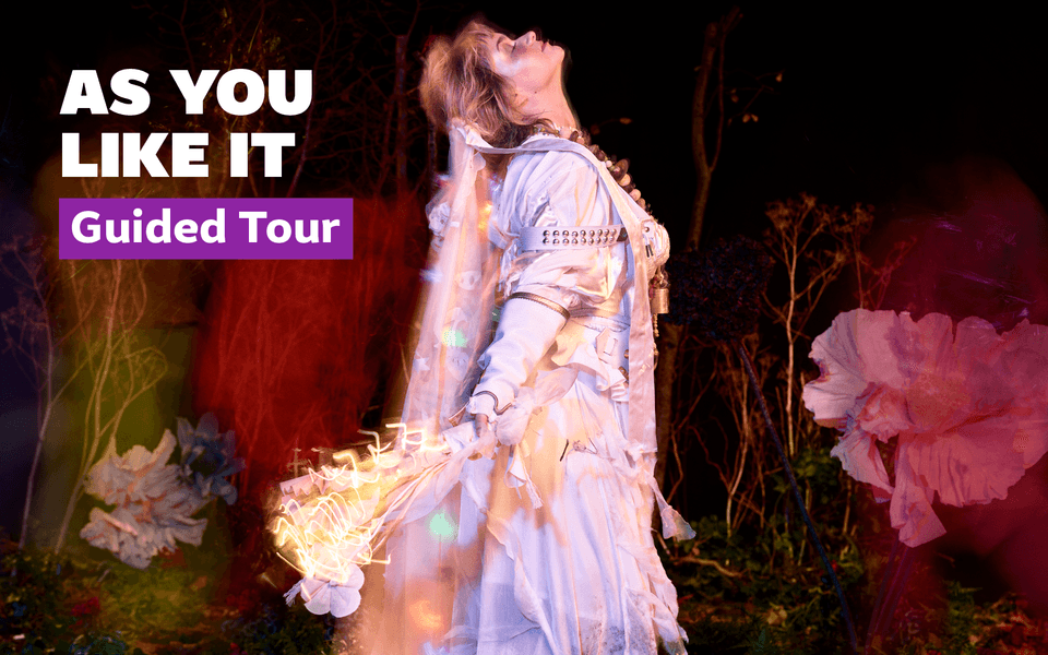 As You Like It. A person wearing white clothes, jewellery and a veil stands in front of a dark forest, with pink flowers around them.
