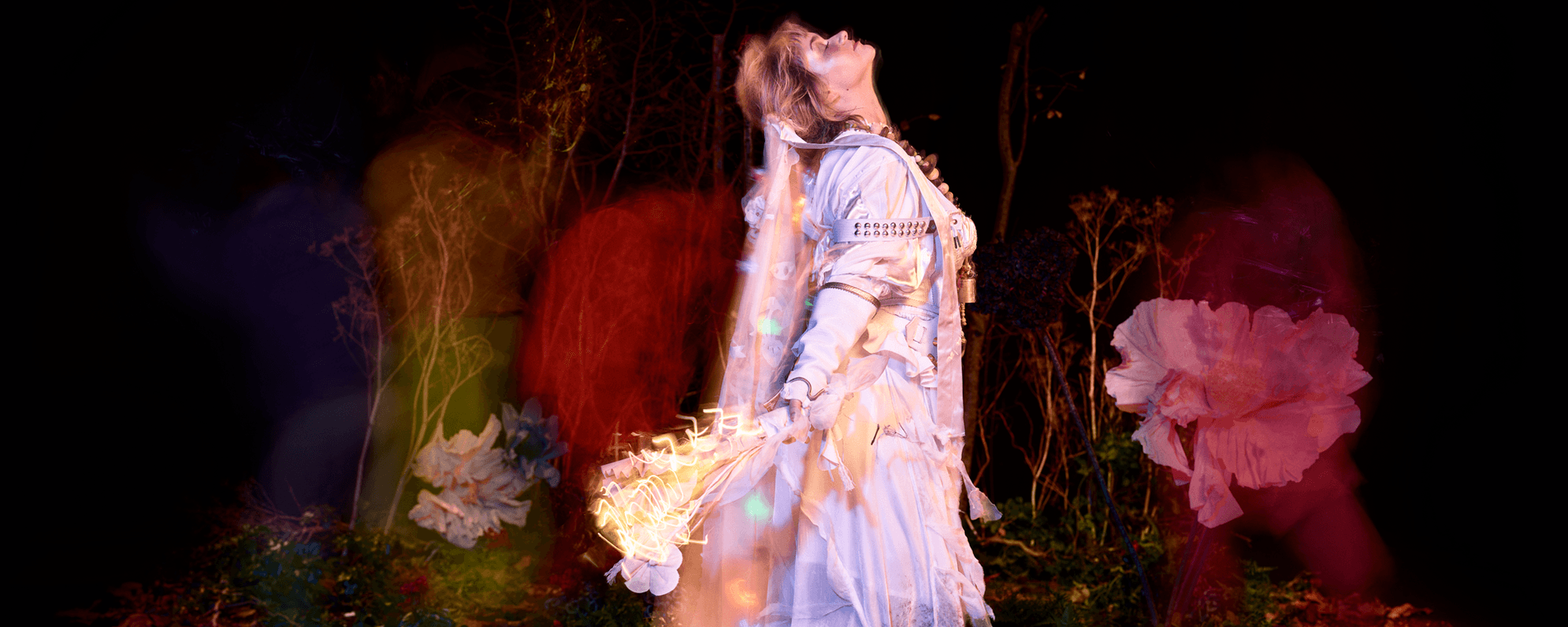 A person wearing a white shirt and a pinafore dress with a veil, smiles gently directly into the camera, whilst pointing up with their right hand. They have blonde hair and brown eyes and wear a large bead necklace in various shades of brown. Small orbs of colour appear around them. In the background, various trees are overshadowed by tints of red, yellow and purple.